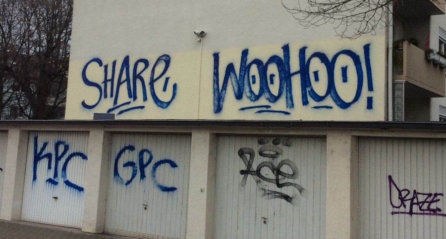 Hier ist ein Bild von einer Hauswand mit Garagen zu sehen. Das englische Graffiti darauf lautet "Share. Wohooo!"