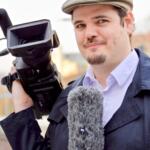 Adrian trägt eine helle Kappe und einen blauen Sommermantel. In der rechten Hand hält er eine kleine Fernsehkamera, in der linken Hand ein Mikrofon mit Windschutz