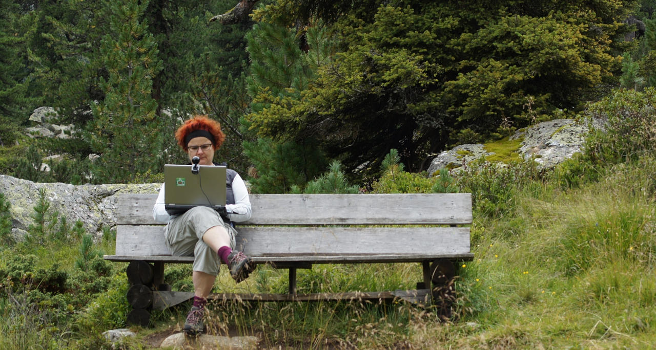 Eine Person sitzt in der Natur mit einem Notebook auf dem Schoß darauf ist eine Kamera angebracht