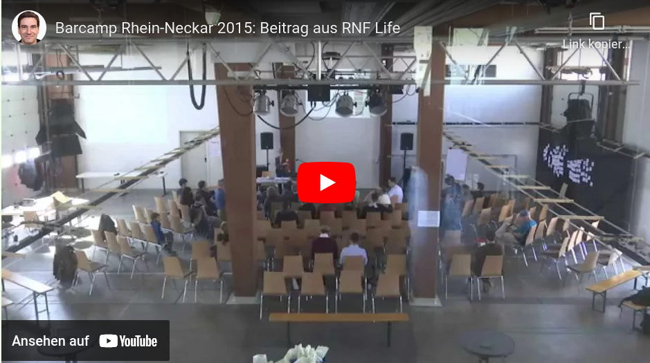 Video-Startbild: Blick auf den Hauptraum der Veranstaltung. Stuhlreihen. Etwa 20 Personen lauschen einer Session.